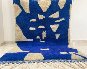 Puede incluir: Una alfombra con estampado geométrico azul y beige. La alfombra presenta un fondo azul intenso con formas beige, incluyendo triángulos y rectángulos. La alfombra tiene flecos a lo largo del borde inferior.