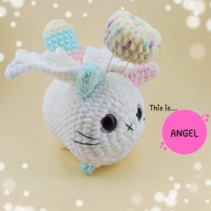 Può includere: Un angelo bianco e morbido, all'uncinetto, con grandi occhi neri e una bocca cucita. L'angelo ha ali e accenti colorati in tonalità pastello di rosa, blu e giallo. Il testo "ANGEL" è in un cerchio rosa.