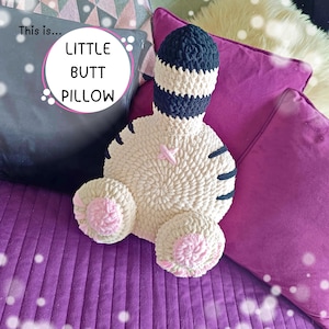 Op de afbeelding: Een handgemaakt, crèmekleurig gehaakt kussen in de vorm van een cartoonkont, met een gestreepte marineblauwe en witte bovenkant en zwarte strepen. Het kussen heeft roze accenten en de tekst "LITTLE BUTT PILLOW".