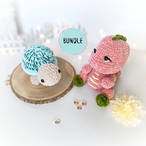 Può includere: Un dinosauro rosa all'uncinetto e una tartaruga azzurra e beige all'uncinetto. Il dinosauro ha foglie verdi sulla testa e piedi verdi. La tartaruga è seduta su una base rotonda di legno. La parola "BUNDLE" è scritta in un cerchio blu chiaro.