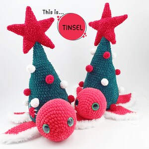 Può includere: Due decorazioni per l'albero di Natale all'uncinetto con stelle rosse in cima. Gli alberi sono verdi con pompon bianchi e rossi. Le decorazioni hanno volti in crochet rosso con occhi verdi. Il testo "This is... TINSEL" è visibile nell'immagine.