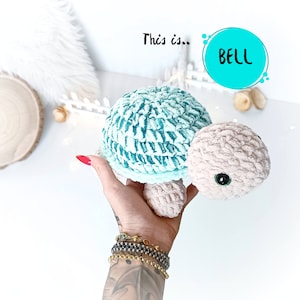Puede incluir: Un juguete de tortuga de crochet con una concha de color verde azulado y blanco y un cuerpo beige. El juguete tiene ojos negros y una expresión amigable. El texto "This is... BELL" es visible en el fondo.