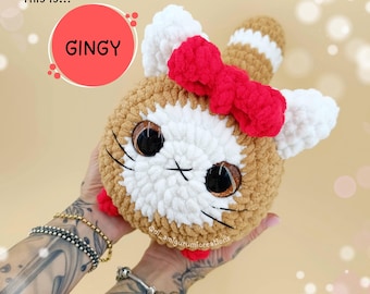 May include: A handmade crochet plush toy in the shape of a cat, with brown, white, and red accents. The cat has large, expressive eyes, a red bow, and the word "GINGY" in a red circle. The toy is held in someone's hands.