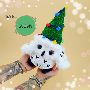 Può includere: Un gufo bianco fatto a mano con un cappello a forma di albero di Natale verde decorato con luci colorate. Il testo "This is... GLOWY" è visibile. Una decorazione festiva per le vacanze.