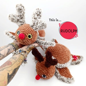 Può includere: Due giocattoli di renne all'uncinetto fatti a mano. Le renne hanno corpi marroni, corna grigie e marroni soffici e nasi rossi. Il testo "This is... RUDOLPH" è in un cerchio rosso.