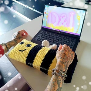 Può includere: Un cuscino fatto a mano a forma di ape a strisce gialle e nere, con un volto sorridente e ali di pompon bianche. Un computer portatile è aperto sullo sfondo, che mostra testo rosa. Il cuscino poggia su una superficie bianca.