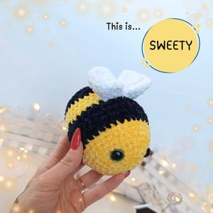 Può includere: Un giocattolo di peluche a forma di ape lavorato a uncinetto con strisce gialle e nere, ali bianche e occhi neri. Il testo "This is... SWEETY" è visibile sullo sfondo.