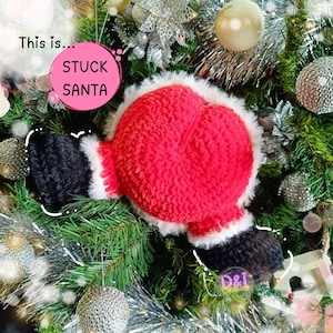 Può includere: Un ornamento per albero di Natale fatto a mano con un corpo rosso di Babbo Natale con finiture bianche e stivali neri. L'ornamento è posto tra aghi di pino verdi e ornamenti argentati e dorati. Il testo "This is... STUCK SANTA" è visibile.