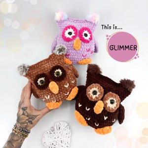 Puede incluir: Tres peluches de búho de crochet. Uno es morado con ojos rosas, otro es marrón con ojos marrones y el último es marrón con ojos marrones. Los búhos están todos sentados en una superficie blanca. Esto es... GLIMMER