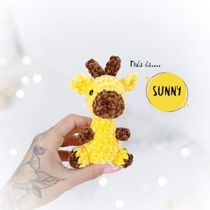 May include: A small yellow crocheted giraffe with brown ears and a brown nose. The giraffe is being held in a hand. The text "This is.... SUNNY" is in the background.