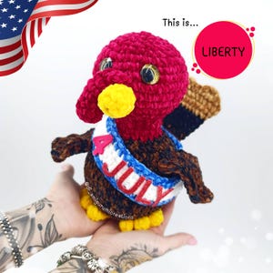 Può includere: Una bambola di tacchino all'uncinetto fatta a mano con testa rossa, corpo marrone e piedi gialli. Il tacchino indossa una fascia blu, bianca e rossa con la scritta "4 JULY". Un cerchio rosso con la parola "LIBERTY" è sullo sfondo.