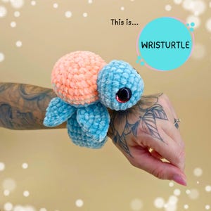 Può includere: Un braccialetto da polso fatto a mano a forma di tartaruga. La tartaruga ha un guscio color pesca, una testa e pinne blu e grandi occhi neri con dettagli rossi. Il testo "WRISTURTLE" è in una nuvoletta.