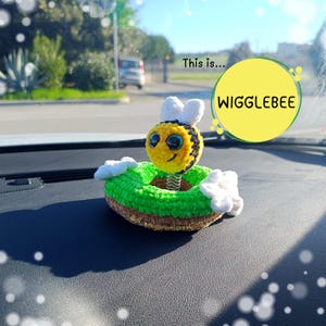 May include: A crocheted bee figure with black and yellow stripes, white wings, and large dark eyes, mounted on a metal spring atop a green and brown crocheted base with white flowers. A yellow circle graphic contains the text This is... WIGGLEBEE.