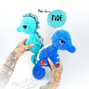 May include: Two crocheted seahorse plushies, one blue and one turquoise, with white trim. The blue seahorse has a red heart on its side. The text "This is... Tide" is written in a white circle above the seahorses.