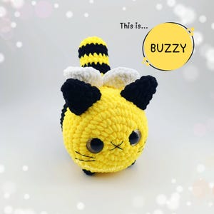 May include: A handmade yellow and black crocheted bee-cat plushie with large, expressive eyes. The plushie has black and white wings, black ears, and a striped tail. The word "BUZZY" is in a yellow circle.