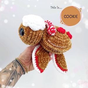 以下が含まれることがあります： 茶色、白、赤の色合いの、かぎ針編みのカメのぬいぐるみ。カメは、頭と足に白いアクセントと赤いディテールを備えた、質感のある豪華なボディをしています。「This is... COOKIE」というテキストが画像にあります。