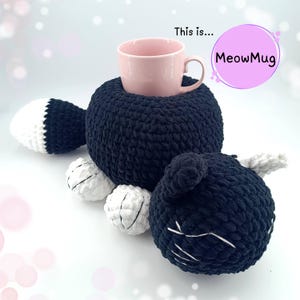 Puede incluir: Un posavasos de ganchillo negro y blanco con forma de gato con una taza rosa en su interior. El posavasos tiene un cuerpo redondo, una cola y una cabeza de gato con bigotes blancos. El texto "MeowMug" está en un círculo rosa.