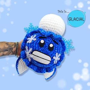 Può includere: Una creatura blu all'uncinetto con dettagli di fiocchi di neve bianchi e una parte superiore bianca. La creatura ha una bocca ampia e sorridente e occhi neri. Il testo "This is... GLACIAL" è sullo sfondo.