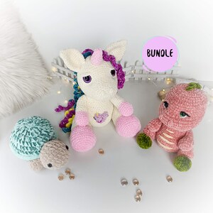 Può includere: Un unicorno all'uncinetto con un corno rosa, una tartaruga a righe rosa e verde e un dinosauro rosa all'uncinetto con una coda verde. L'unicorno ha un cuore rosa sul petto. La parola "BUNDLE" è scritta in rosa su un cerchio bianco.