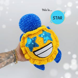 Può includere: Una tartaruga di peluche blu e gialla fatta a mano con un volto sorridente e occhi a forma di stella. Il guscio è giallo con occhi a stella di paillettes blu e una bocca bianca. Il testo "STAR" è in un cerchio blu.