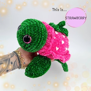 Può includere: Un giocattolo di peluche di tartaruga all'uncinetto verde e rosa con occhi grandi. Il guscio della tartaruga è rosa con cuori bianchi e il testo "STRAWBERRY" è scritto in un cerchio rosa.