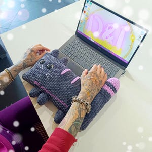Könnte beinhalten: Ein dunkelgraues, gehäkeltes Laptop-Kissen in Katzenform mit rosa Streifen und Details. Das Kissen liegt auf einem Laptop, mit einer passenden Handgelenkauflage. Der Laptop-Bildschirm zeigt eine farbenfrohe Grafik. Das Produkt ist handgefertigt.