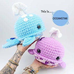 May include: Two crocheted whale plushies, one light blue with white spots and a white fin, the other purple with white and purple stripes and a white fin. The text "This is... OCEANSTAR" is in the background.