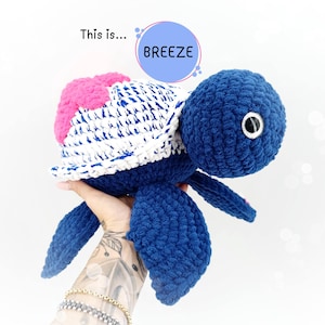 Può includere: Una tartaruga all'uncinetto blu con un guscio a strisce bianche e blu e accenti rosa. La tartaruga ha un grande occhio rotondo ed è tenuta da una mano.
