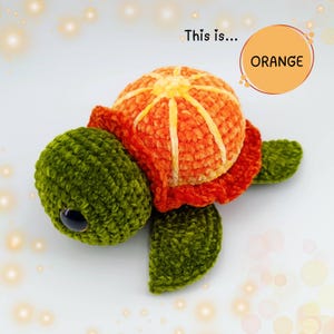 Può includere: Un giocattolo di peluche a forma di tartaruga all'uncinetto con un guscio arancione e un corpo verde. Il guscio è progettato per assomigliare a un'arancia con un design segmentato. Il testo "This is... ORANGE" è visibile nell'immagine.