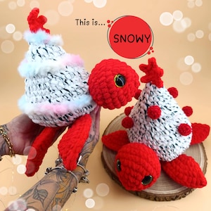 Può includere: Due giocattoli di tartaruga all'uncinetto con corpi rossi e gusci bianchi e neri. I gusci sono decorati con pelliccia bianca e pompon rossi. Il testo "This is... SNOWY" è visibile sullo sfondo.
