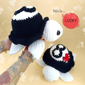 Puede incluir: Dos peluches de tortuga de crochet, uno negro y blanco con una pajarita negra y el otro negro y blanco con un corazón rojo en el caparazón. El texto "This is... LUCKY" está en un círculo rojo.