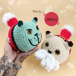 May include: Two crocheted cat toys wearing Santa hats. The cat on the left is green and the cat on the right is beige. The text "This is... JINGLE" is in the background.