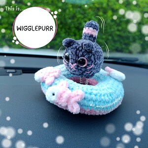 Può includere: Una figura di gatto all'uncinetto grigio scuro con grandi occhi neri e naso ricamato rosa, montata su una molla sopra un anello all'uncinetto azzurro e rosa decorato con piccoli pesci rosa. Il testo "This is... WIGGLEPURR" è presente nell'immagine.