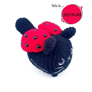 May include: A black crocheted cat-shaped plush toy with a red bow featuring black spots. The toy has large, round eyes and white stitched details. The text "LADYPURR" is in a red circle.