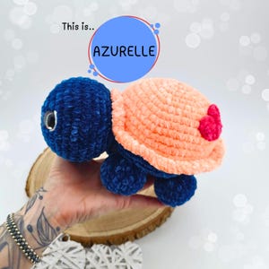May include: A crocheted plush turtle toy with a blue body, peach-colored shell, and pink heart accents. The toy is held in a hand.