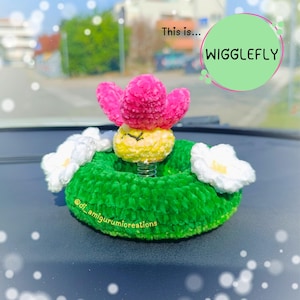 Può includere: Una base ad anello verde all'uncinetto con due fiori bianchi, caratterizzata da una creatura gialla e rosa su una molla metallica al centro. Il testo "This is... WIGGLEFLY" appare in un cerchio verde e "@di_amigurumicreations" è scritto sulla base.