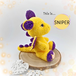 Può includere: Un drago di uncinetto giallo con orecchie, ali e piedi viola. Il drago è seduto su una superficie di legno con il testo "This is... SNIPER" in un cerchio.
