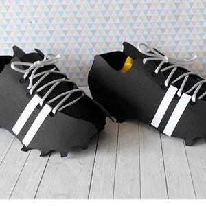 Puede incluir: Un par de botas de fútbol negras con rayas y cordones blancos. Las botas tienen un diseño clásico con una suela texturizada y se exhiben sobre una superficie clara. Los zapatos están hechos de un material mate.