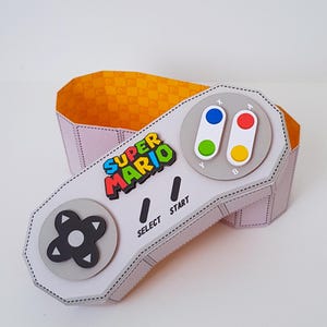 May include: A light pink, paper-crafted box shaped like a classic video game controller. The controller features the Super Mario logo, directional pad, buttons, and the words "SELECT" and "START". The interior is orange with a repeating pattern.