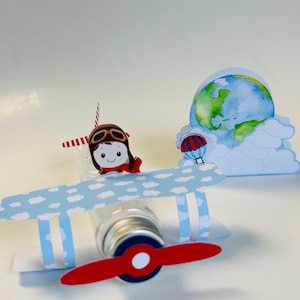 May include: A whimsical airplane-themed party decoration. The centerpiece is a clear bottle adorned with a red and white propeller, blue wings with cloud cutouts, and a cartoon pilot. A globe and hot air balloon complete the travel theme.