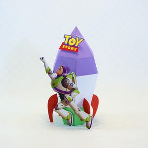 May include: A papercraft Buzz Lightyear figure in a dynamic pose, with a rocket ship backdrop. The rocket is primarily white, purple, and red, with the "Toy Story" logo at the top. Buzz Lightyear is in his signature green, white, and purple spacesuit.