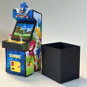 May include: A small arcade machine decorated with Sonic the Hedgehog characters. The machine is predominantly blue and black, with the word "SONIC" visible. A black box is positioned next to the arcade machine.