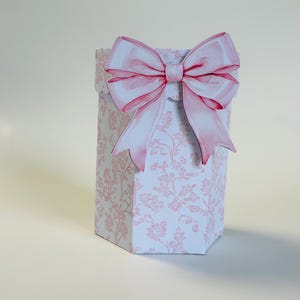 May include: A decorative gift box with a large pink bow. The box is white with a pink floral pattern. The bow is a watercolor style with pink and white hues. The box is hexagonal in shape.
