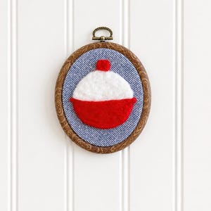 May include: An oval-shaped, framed wall hanging featuring a red and white bobber design. The bobber is set against a blue denim-like background, framed by a wooden oval. A small metal loop is at the top for hanging. The wall decor is against a white background.