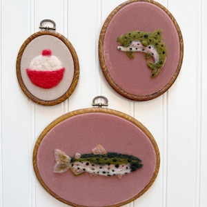 May include: Three framed fiber art pieces. One features a red and white bobber, the other two depict fish. The fish are green, black, and white, set against a pink background. Each piece is in a wooden oval frame with a metal hanger.