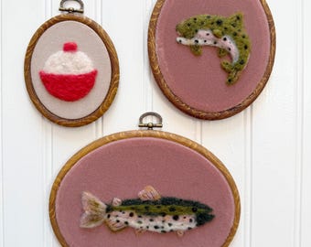 May include: Three framed fiber art pieces. One features a red and white bobber, the other two depict fish. The fish are green, black, and white, set against a pink background. Each piece is in a wooden oval frame with a metal hanger.