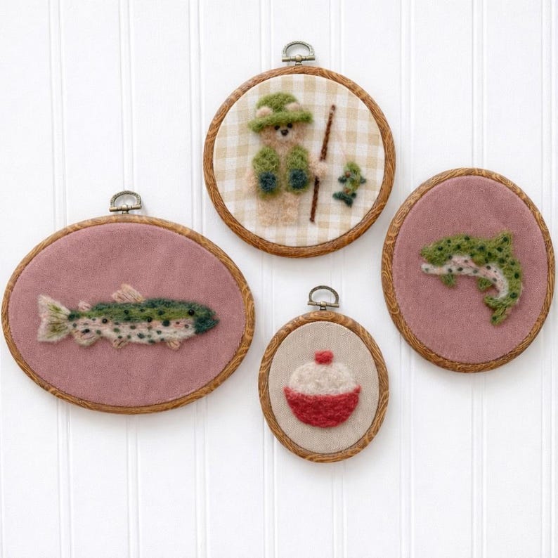 May include: Four framed felt art pieces with fishing themes. The pieces include a fish, a fishing bear, a fish, and a bobber. The frames are wood with a pink or checkered background. Each piece has a metal loop for hanging.