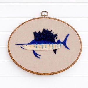 May include: Embroidered sailfish art in a wooden hoop. The fish is blue with a white and light blue belly, and a dark blue dorsal fin. The background is a light beige fabric. The hoop has a brass-colored metal loop at the top for hanging.
