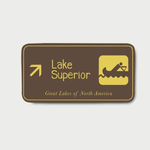 May include: A rectangular brown sign with a yellow border featuring an arrow, the text Lake Superior, and a yellow icon of a person paddling a canoe. Below, the text Great Lakes of North America is printed in a smaller yellow font.