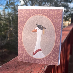 May include: A greeting card featuring a goose wearing a graduation cap and a red sash. The goose is inside an oval frame with a floral design. The card is set against a blurred background of trees and a red wooden surface.